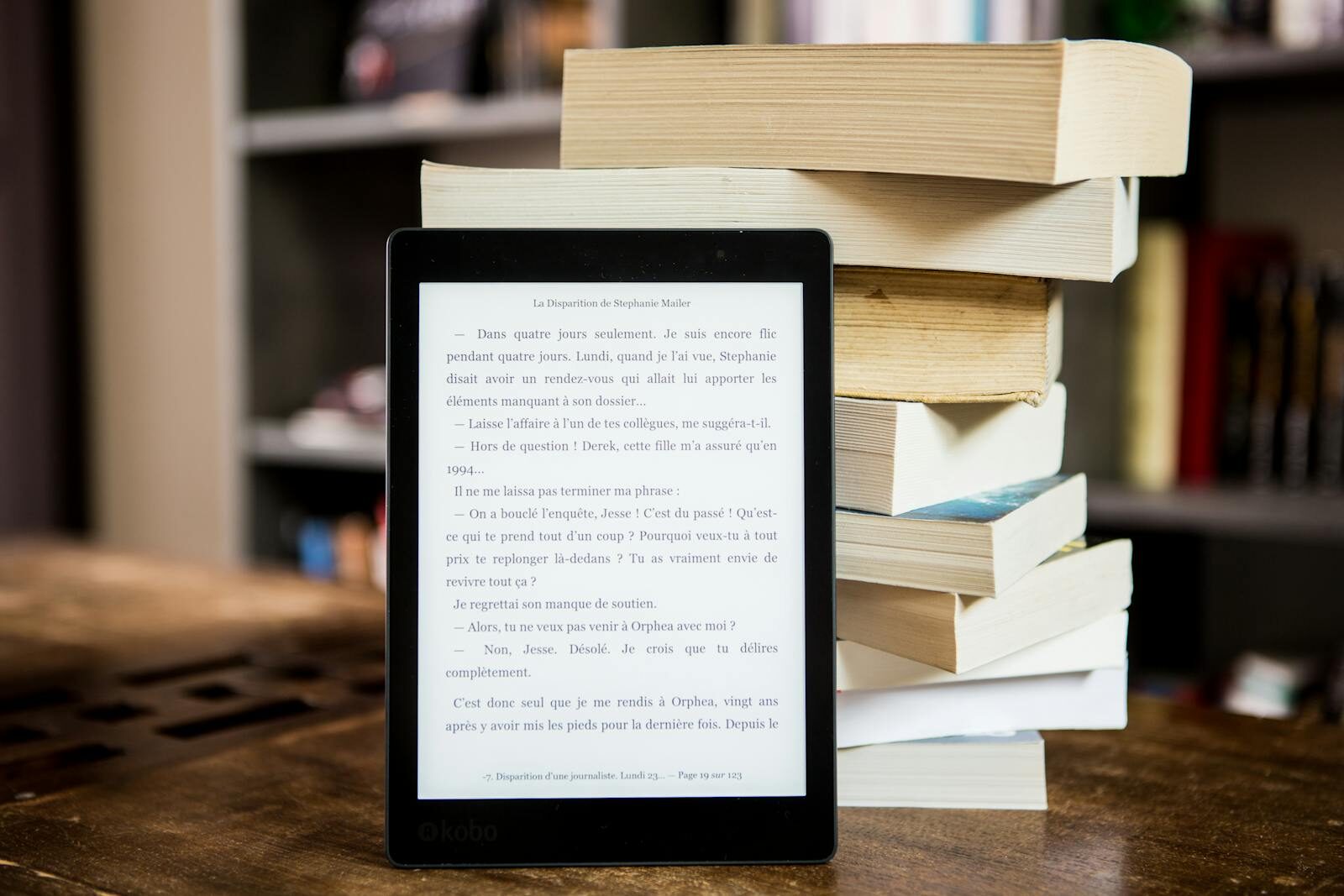 E-reader displaying text from "La Disparition de Stephanie Mailer," beside a stack of various books, highlighting a blend of digital and traditional reading.