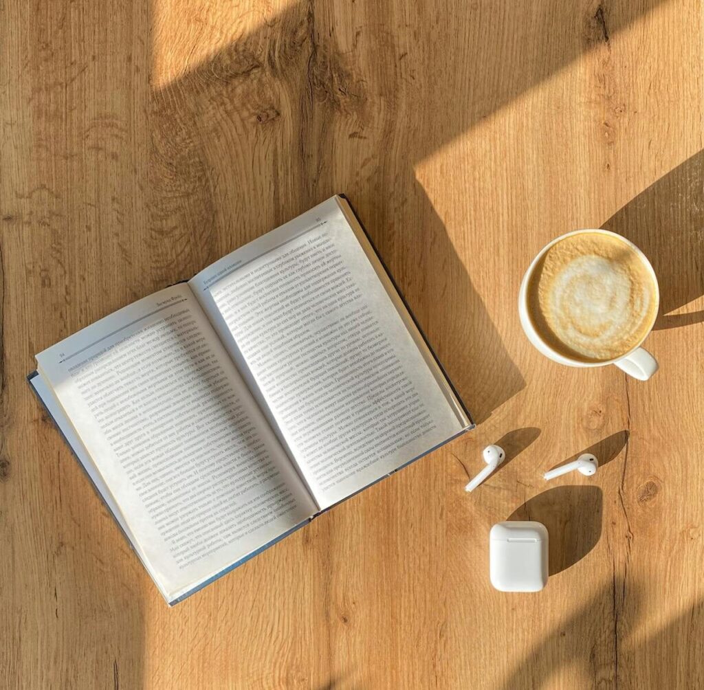 Open book on a wooden table next to a latte and wireless earbuds, suggesting a cozy reading or study session.