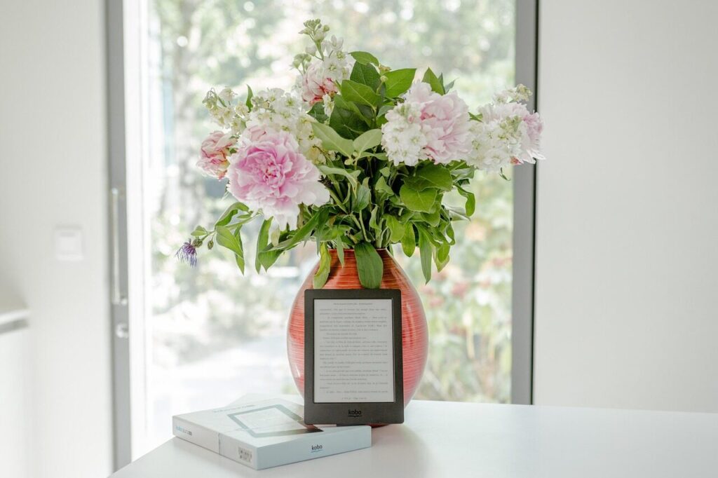 A colorful vase filled with pink and white flowers sits beside a Kobo e-reader on a white surface, highlighting a cozy reading space.