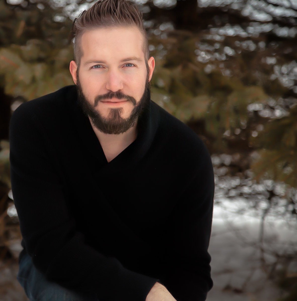 A man with a beard poses outdoors in a black sweater, surrounded by snow and evergreen trees, conveying a serene winter atmosphere.