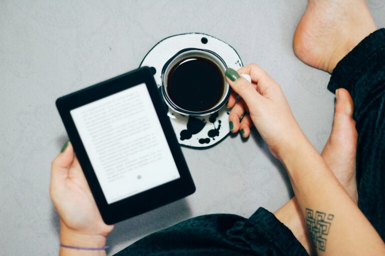 A person holds an e-reader in one hand while sipping coffee from a patterned cup, seated comfortably on a soft surface.