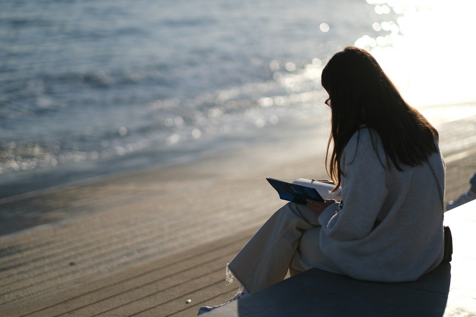 A person reading a book by the water, sunlight reflecting off the waves. The scene conveys a peaceful moment of leisure and relaxation.