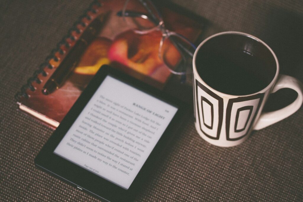 A cozy reading setup featuring an e-reader displaying text, a patterned coffee mug, a notebook, and glasses, suggesting a relaxed study atmosphere.